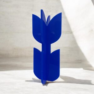 A neon acrylic flower sculpture stands at the center of the image. The object consists of two interlocking pieces that form four smooth, petal-like shapes arranged in a cross. The glossy, translucent material glows brightly against the grayscale background. Abstract geometric shapes and fine vertical lines create a modern, sculptural, almost futuristic setting. The vivid neon object becomes the clear visual focus, radiating color and contemporary design.