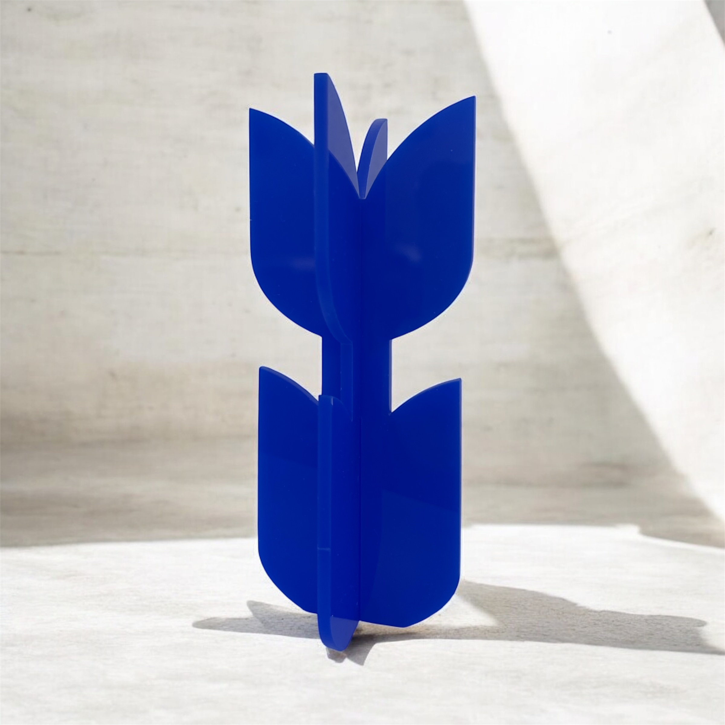 A neon acrylic flower sculpture stands at the center of the image. The object consists of two interlocking pieces that form four smooth, petal-like shapes arranged in a cross. The glossy, translucent material glows brightly against the grayscale background. Abstract geometric shapes and fine vertical lines create a modern, sculptural, almost futuristic setting. The vivid neon object becomes the clear visual focus, radiating color and contemporary design.