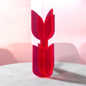 A neon acrylic flower sculpture stands at the center of the image. The object consists of two interlocking pieces that form four smooth, petal-like shapes arranged in a cross. The glossy, translucent material glows brightly against the grayscale background. Abstract geometric shapes and fine vertical lines create a modern, sculptural, almost futuristic setting. The vivid neon object becomes the clear visual focus, radiating color and contemporary design.