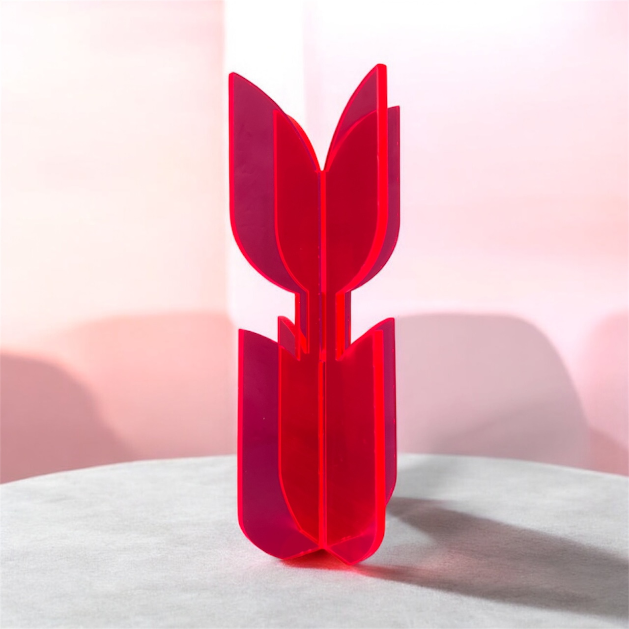A neon acrylic flower sculpture stands at the center of the image. The object consists of two interlocking pieces that form four smooth, petal-like shapes arranged in a cross. The glossy, translucent material glows brightly against the grayscale background. Abstract geometric shapes and fine vertical lines create a modern, sculptural, almost futuristic setting. The vivid neon object becomes the clear visual focus, radiating color and contemporary design.
